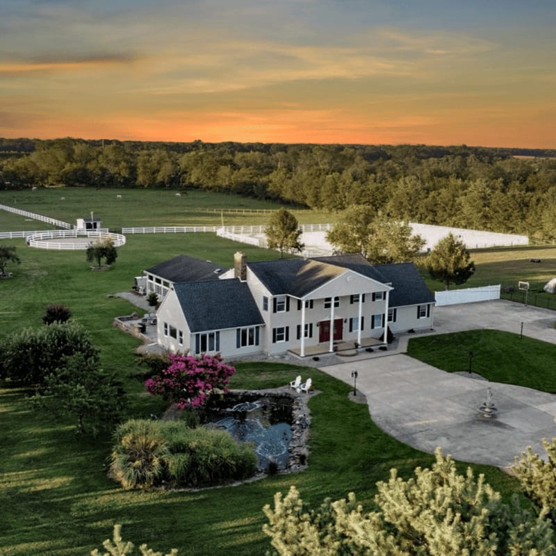 A grand estate with a pond and lush landscaping in the foreground is set against a backdrop of rolling hills and a vibrant sunset.