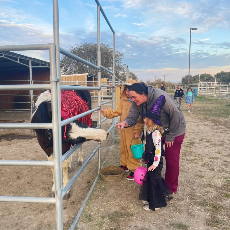 A woman and two children in costumes interact with a cow in a pen, with a rural landscape and a few other people visible in the background.