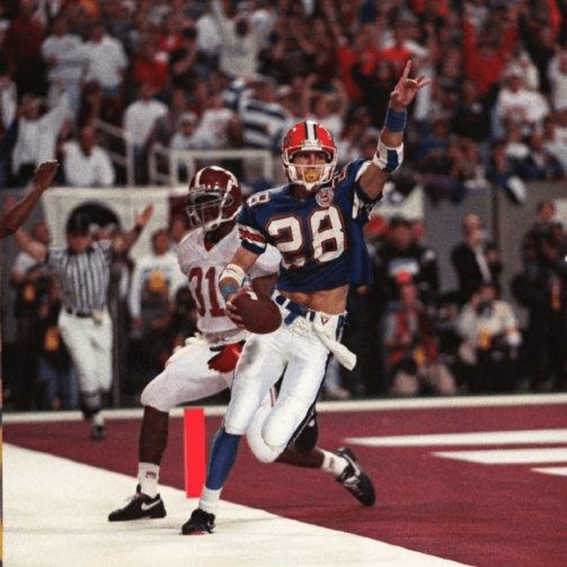 A football player in a blue jersey with the number 28 celebrates a touchdown in the foreground, while a blurred crowd cheers in the background.