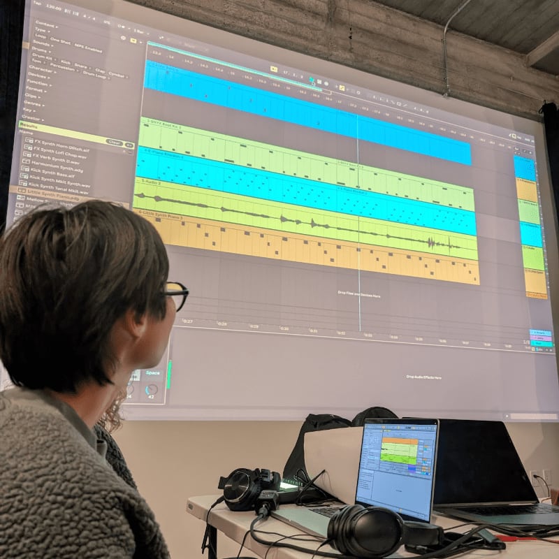 A person wearing glasses and a fuzzy jacket looks at a large projection screen displaying a music production software interface, with a laptop and headphones on a table in the foreground.