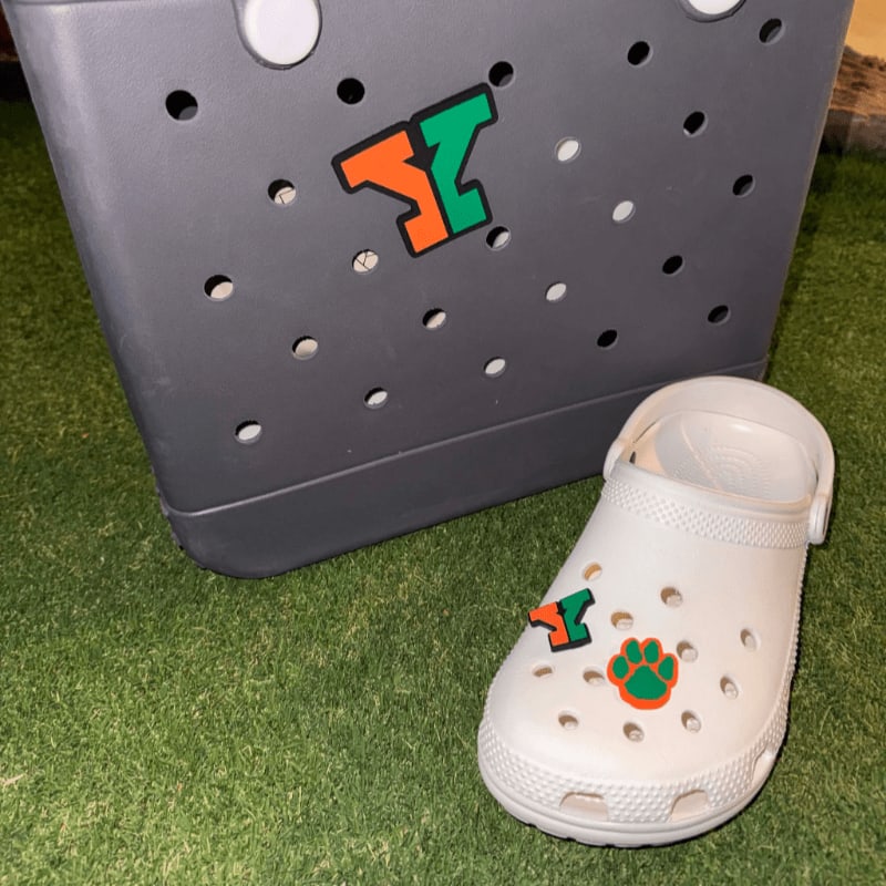 A white Croc with an orange and green paw print charm sits on a green surface in front of a gray bag adorned with a matching orange and green "Y" charm.