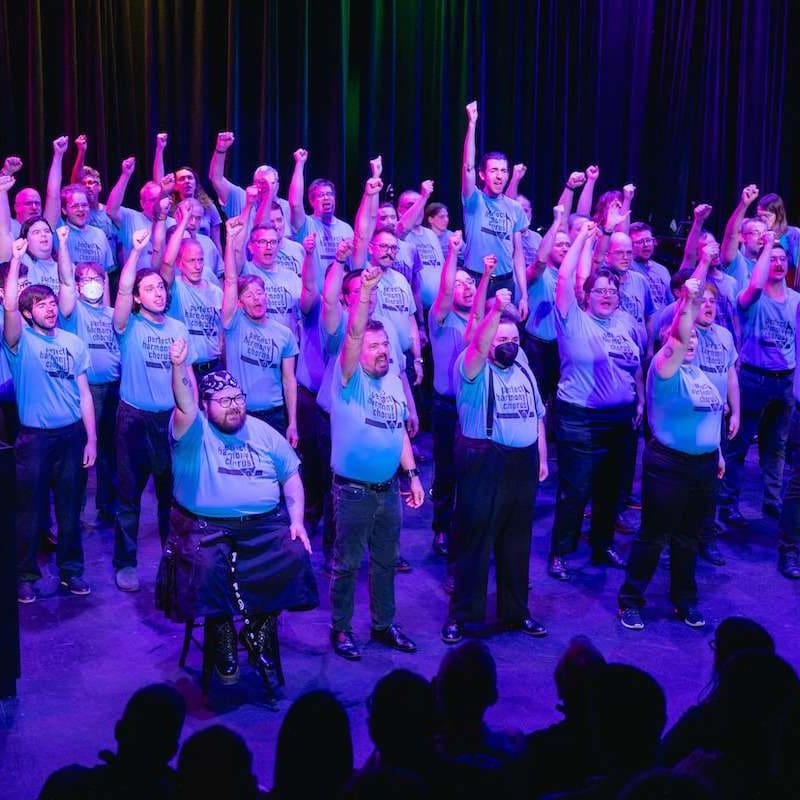 A choir of men in blue t-shirts with fists raised stands on a stage with a colorful curtain in the background.