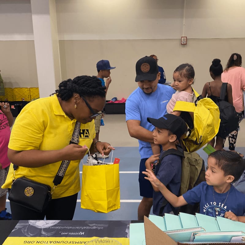 A woman in a yellow shirt hands a yellow bag to a boy in a blue shirt, while a man holds a baby and other children look on in the background.