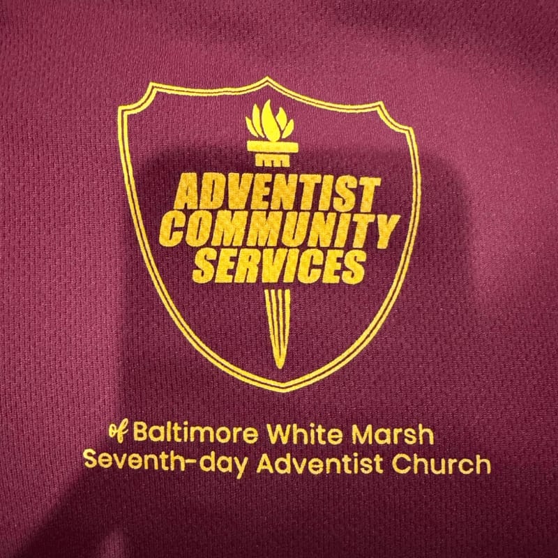 A maroon shirt displays a yellow crest with "ADVENTIST COMMUNITY SERVICES" in the foreground, and "of Baltimore White Marsh Seventh-day Adventist Church" below it in the background.