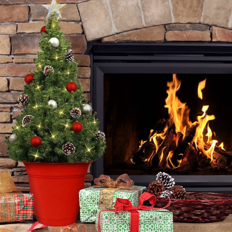 A decorated Christmas tree in a red pot stands in front of a roaring fireplace, with wrapped presents and pinecones scattered on the hearth.