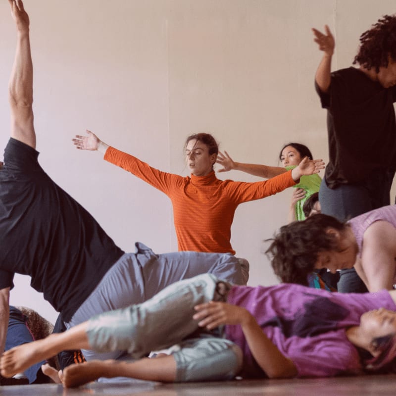 Dancers in various poses occupy the foreground, while a light-colored wall forms the background.