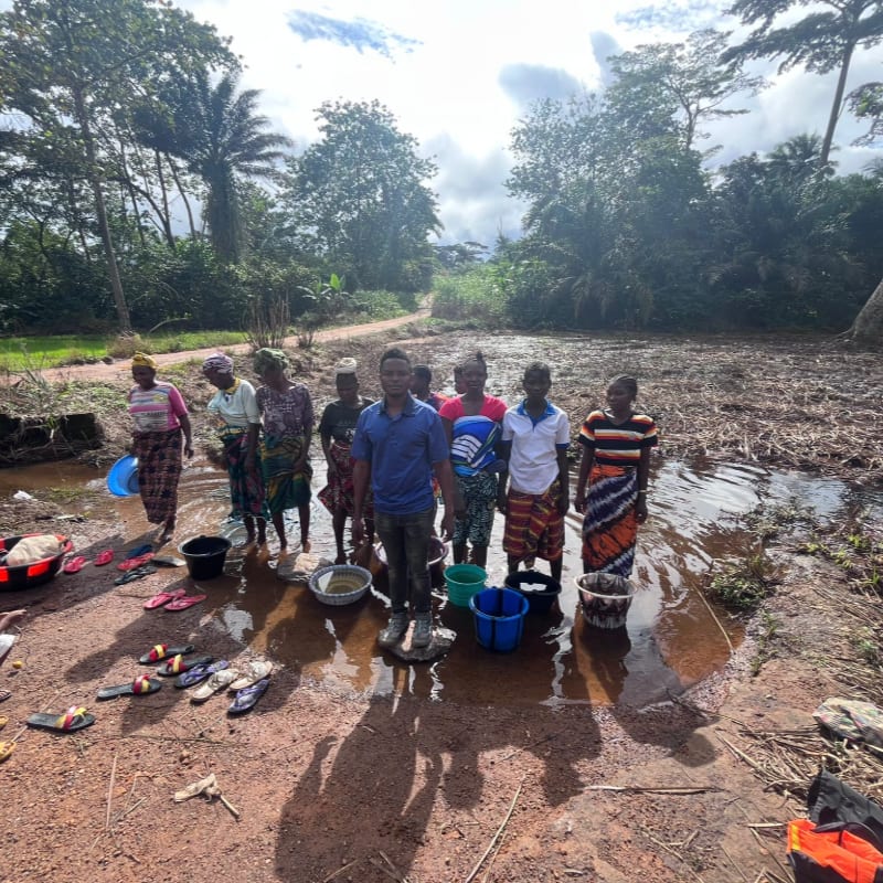 A group of people, including children, stand in a muddy area with buckets and basins, while lush green trees and a cloudy sky form the background.