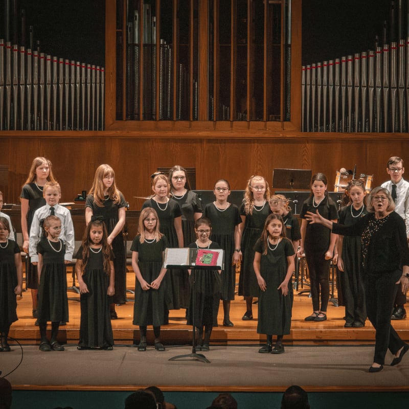 A choir of children in black dresses stands on a stage in front of a large pipe organ.