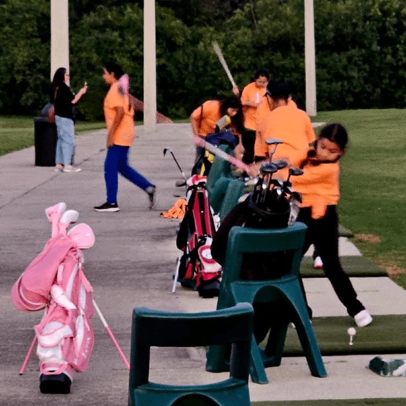 In the foreground, a pink golf bag stands ready, while in the background, several people in orange shirts practice their swings at a driving range.