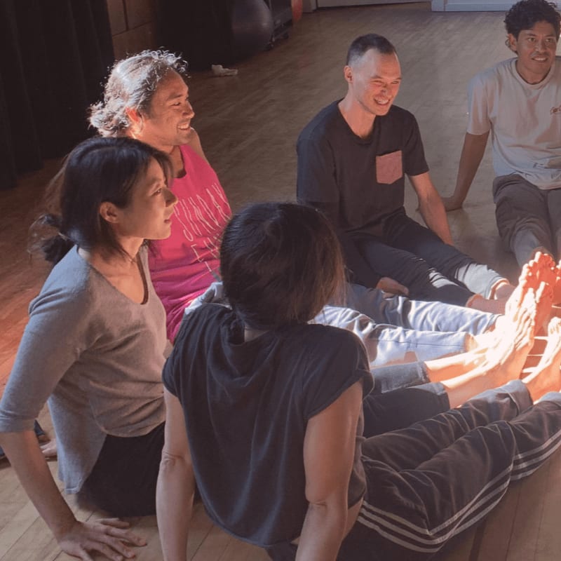 A group of people sit in a circle on a wooden floor, with sunlight streaming in from the right, illuminating their feet.