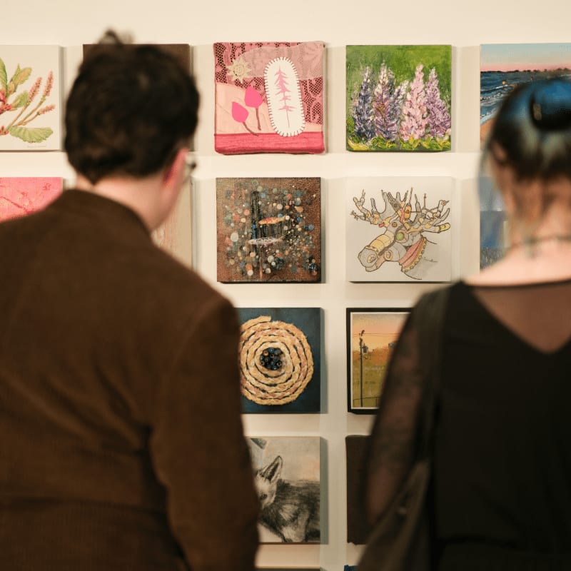 Two people in the foreground are looking at a wall of small, diverse artworks in the background.