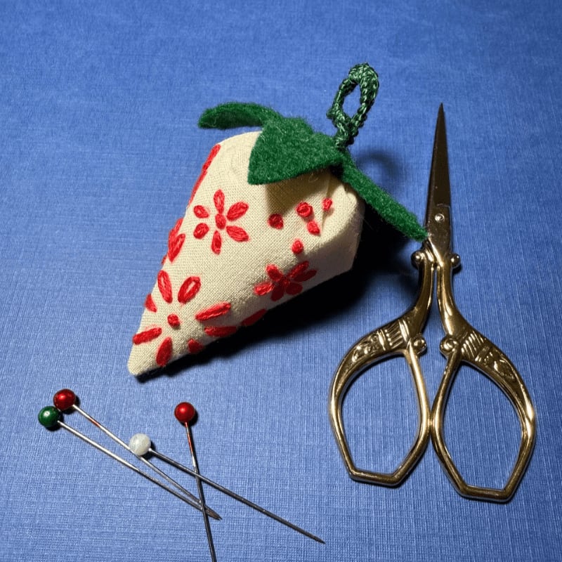 A fabric strawberry pincushion with red floral embroidery sits next to ornate gold scissors and three colorful pins on a blue textured surface.