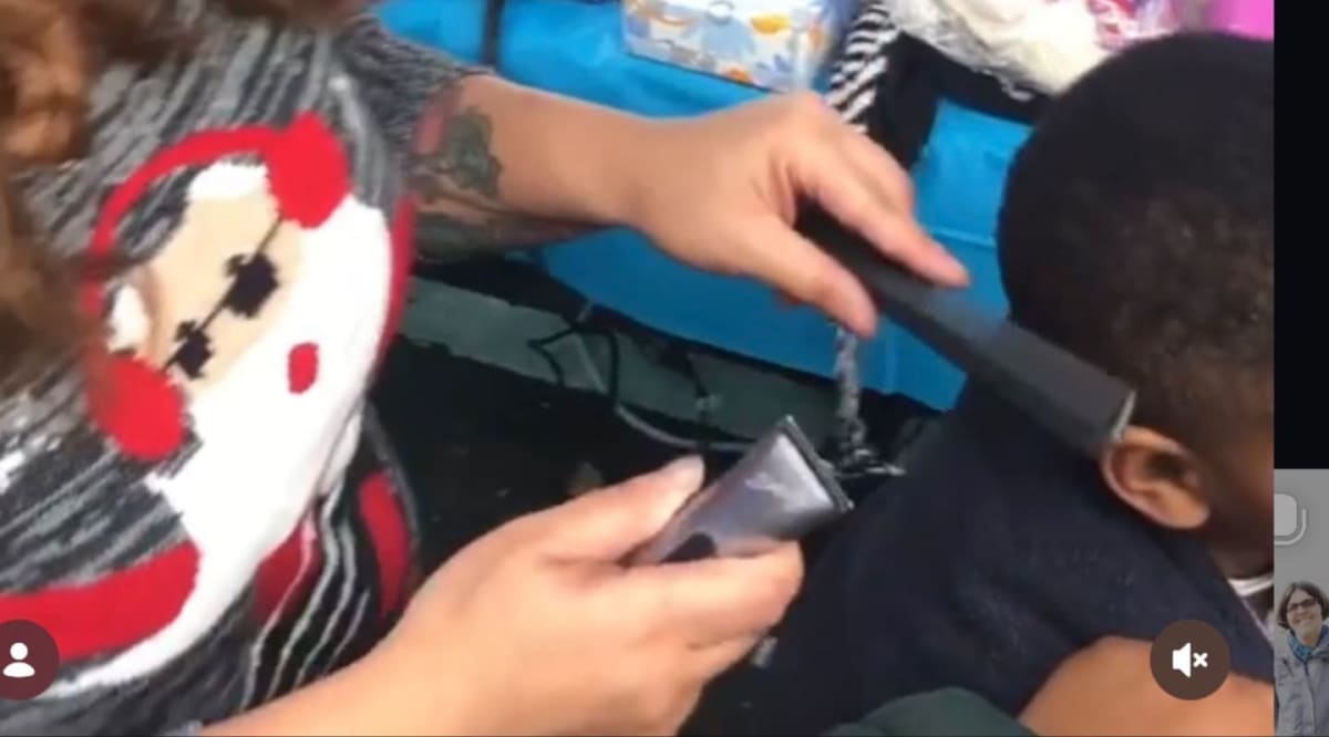 A person with a Santa-themed shirt uses clippers to cut a child's hair, with a blue tablecloth and various items visible in the background.