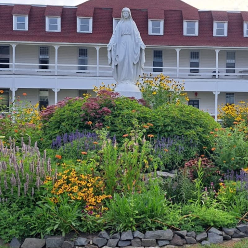 A white statue of Mary stands in a vibrant garden of colorful flowers, with a large white building with a red roof in the background.