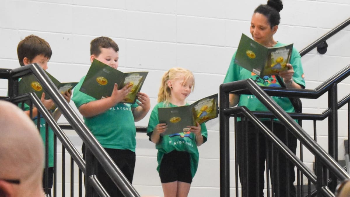 Children and an adult stand on stairs, reading from green books, with a white wall and black railings in the background.