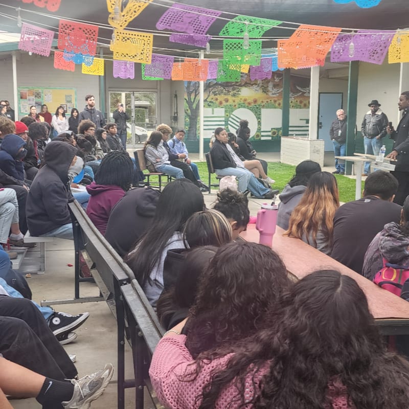 A group of students are seated in the foreground, facing a speaker in the background, with colorful papel picado banners strung overhead.
