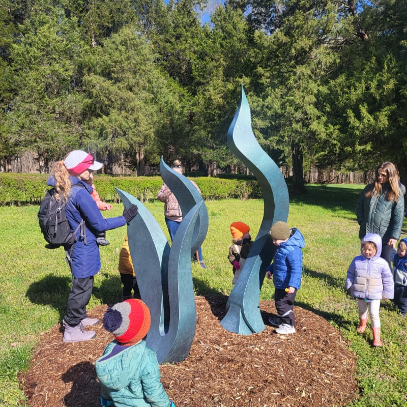 Children in colorful winter clothing gather around a teal, flame-like sculpture in a grassy park with trees in the background.