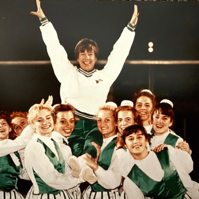 A group of smiling young women in green and white uniforms are cheering, with one woman being lifted into the air in the foreground against a dark background.