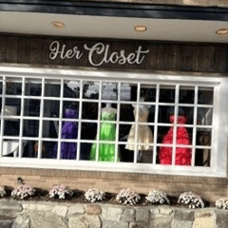 A storefront window displays colorful dresses on mannequins, with a stone facade and small floral arrangements in the foreground.