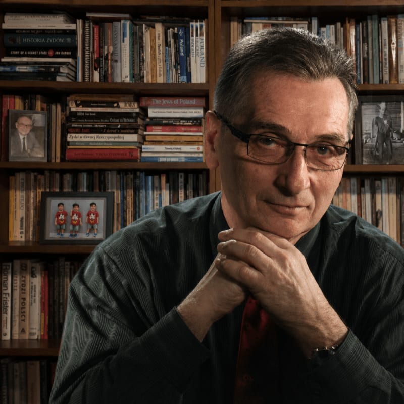 A man with glasses rests his chin on his hands in the foreground, with bookshelves filled with books and framed pictures in the background.