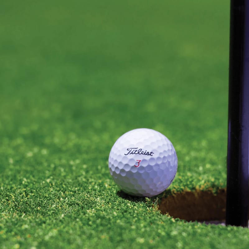 A white Titleist golf ball rests on a vibrant green putting green next to a dark hole, with a blurred green background.