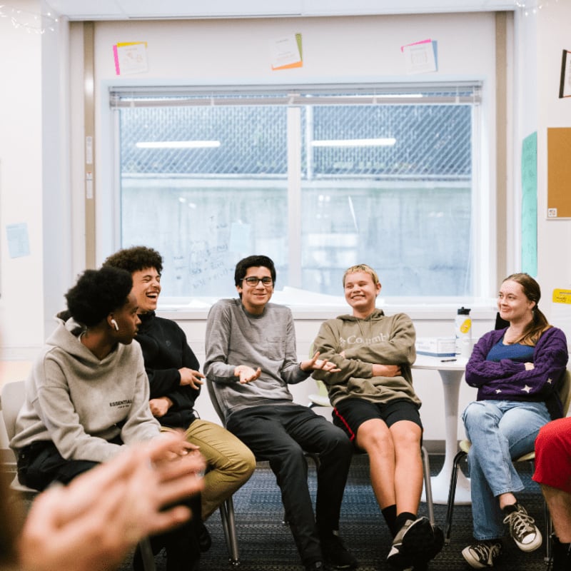 A group of young people are sitting in a circle in a classroom, laughing and talking, with a window and bulletin boards visible in the background.