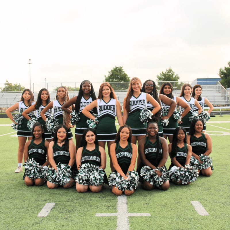 A group of cheerleaders in green and white uniforms pose on a football field with bleachers in the background.