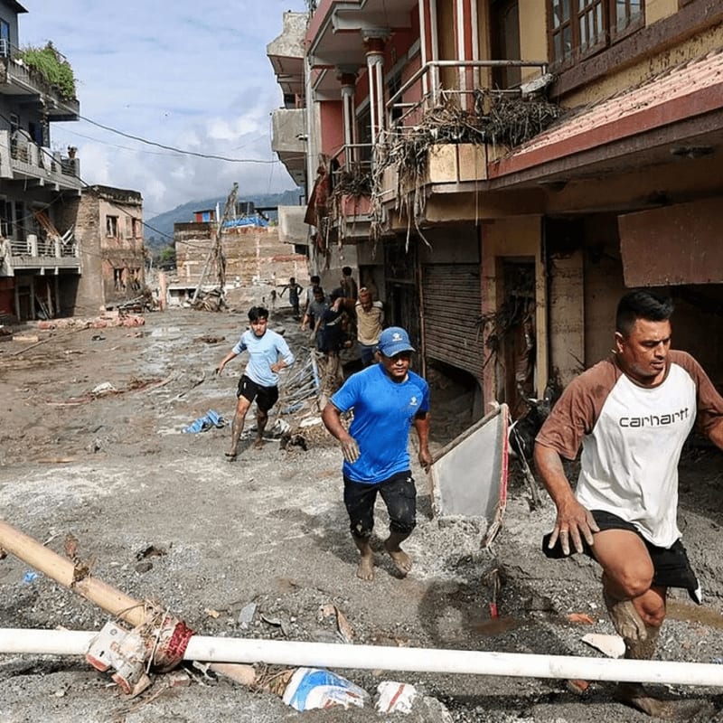 Men run through debris-strewn streets in front of damaged buildings under a cloudy sky.