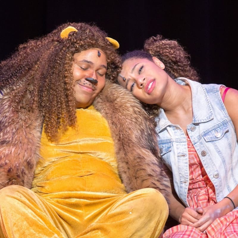 A person in a lion costume rests their head on a young woman's shoulder against a dark stage background.