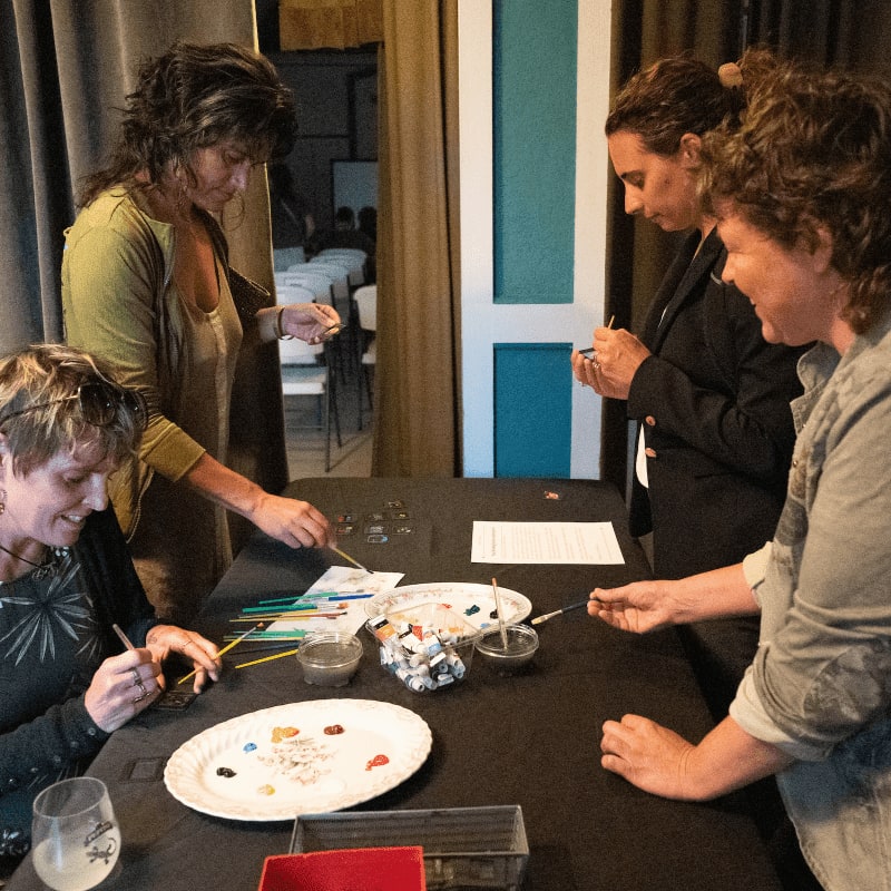 Four women are gathered around a table with art supplies in the foreground, while a blurred background shows a room with curtains and a doorway.