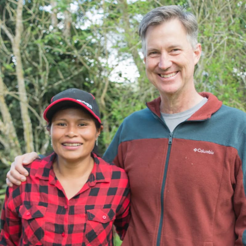 A man and a woman smile at the camera, with lush green foliage in the background.