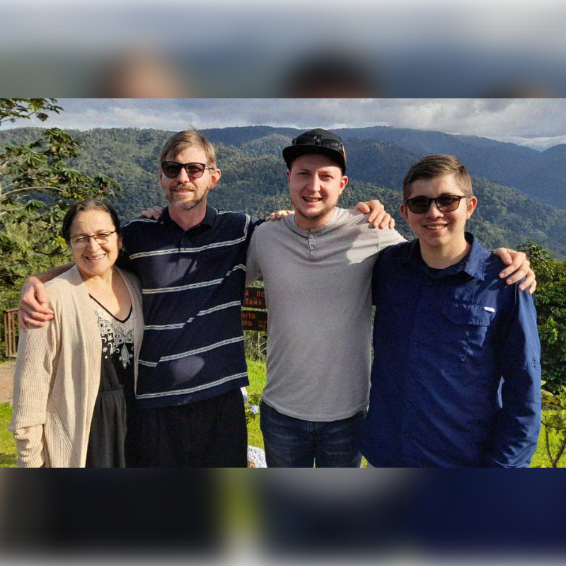 Four people stand with arms around each other in the foreground, with a lush, mountainous landscape in the background.