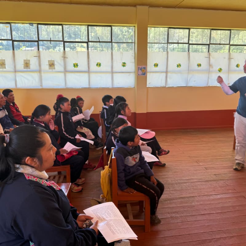 Children and adults sit in a classroom with papers in their hands, while a man stands at the front gesturing towards a window with a bright sky.