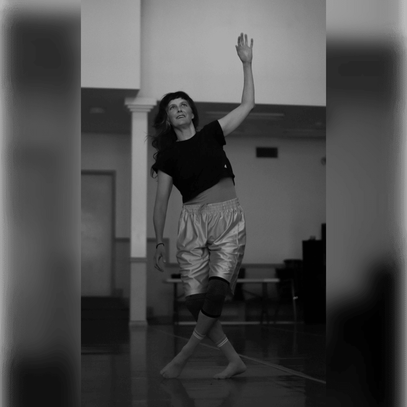 A woman in athletic wear dances with her arm raised in a room with tables and chairs in the background.