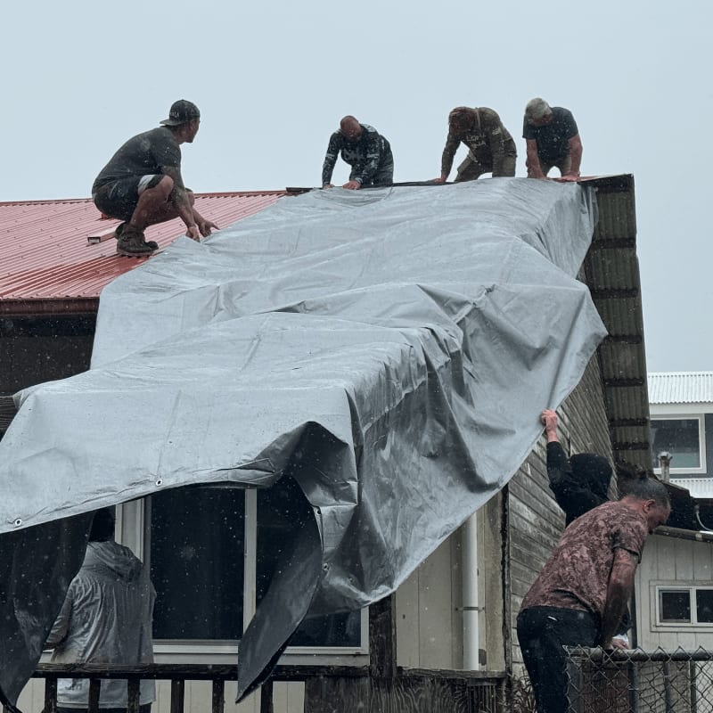 Several people work together on a rooftop in the foreground to secure a large tarp, while a cloudy, overcast sky forms the background.