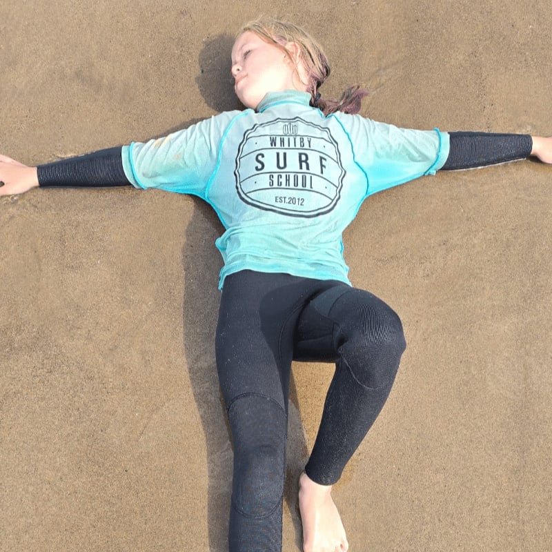 A young girl in a wetsuit lies on the wet sand, with the ocean blurred in the background.