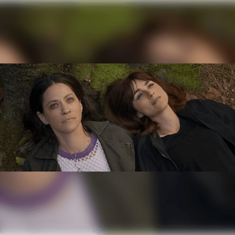 Two women lie on the ground in the foreground, with a blurred background of trees and foliage.