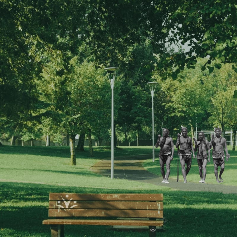 A wooden bench sits in the foreground of a park with a paved path leading into the background where four figures walk under the shade of lush green trees.