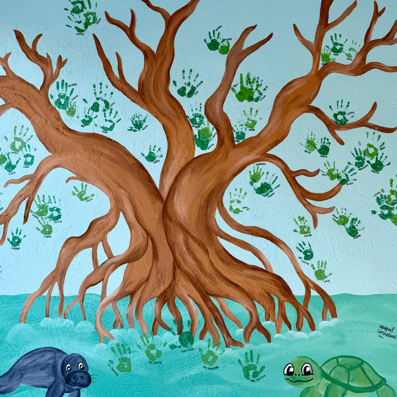 A large brown tree with many green handprints on its branches stands in the foreground, while a manatee and a turtle swim in the turquoise water of the background.