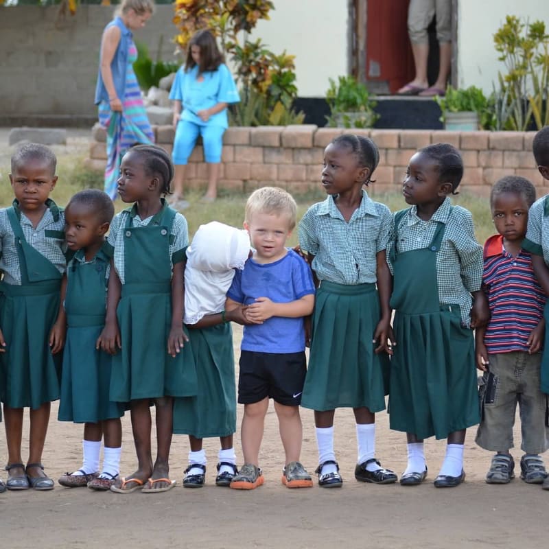A group of young African children in school uniforms stand in the foreground, while adults are visible in the blurred background.