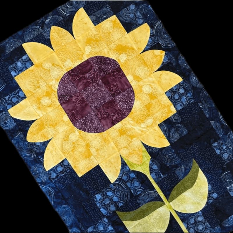 A vibrant yellow sunflower quilt with a dark purple center is set against a dark blue background with swirling patterns.
