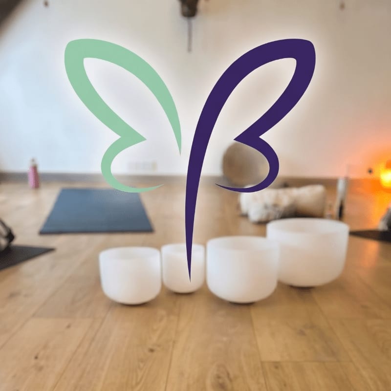A colorful butterfly-like logo hovers over a collection of crystal singing bowls in the foreground, with a yoga mat and soft lighting visible in the blurred background.