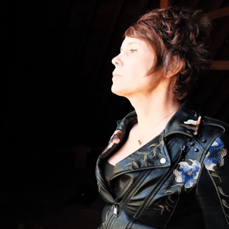 A woman with short, dark hair, wearing a black leather jacket with floral embroidery, looks up and to the left against a dark, shadowy background.