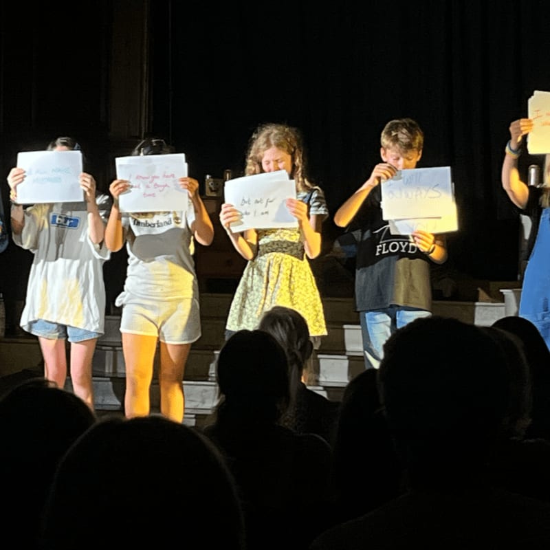 Children stand on a stage holding signs, with an audience visible in the dark foreground.