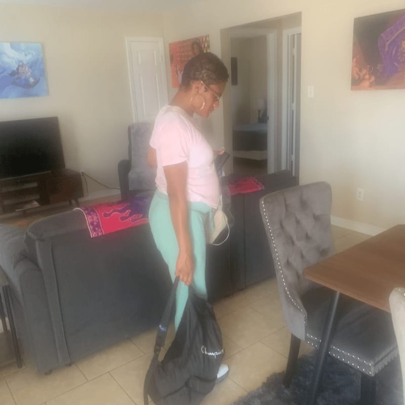 A woman in a pink shirt and teal leggings stands in a living room with a couch and chairs, while artwork hangs on the walls in the background.