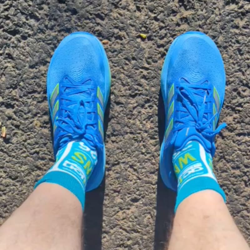 Bright blue running shoes with yellow accents and white socks with green lettering are worn by a person standing on a textured asphalt surface.