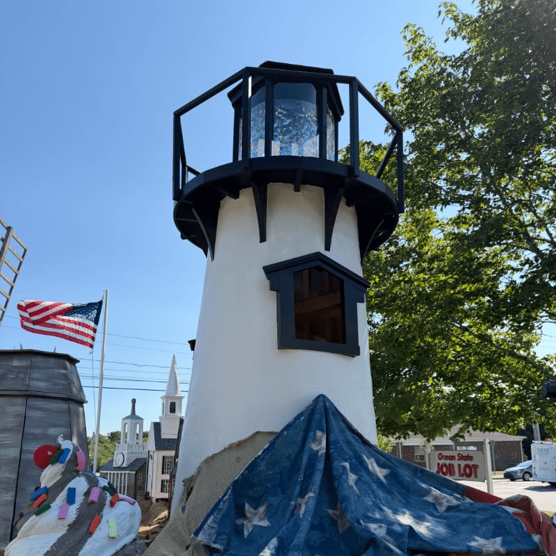 A white lighthouse with a black lantern room stands in the foreground, adorned with a draped American flag, while a clear blue sky and lush green trees form the background.