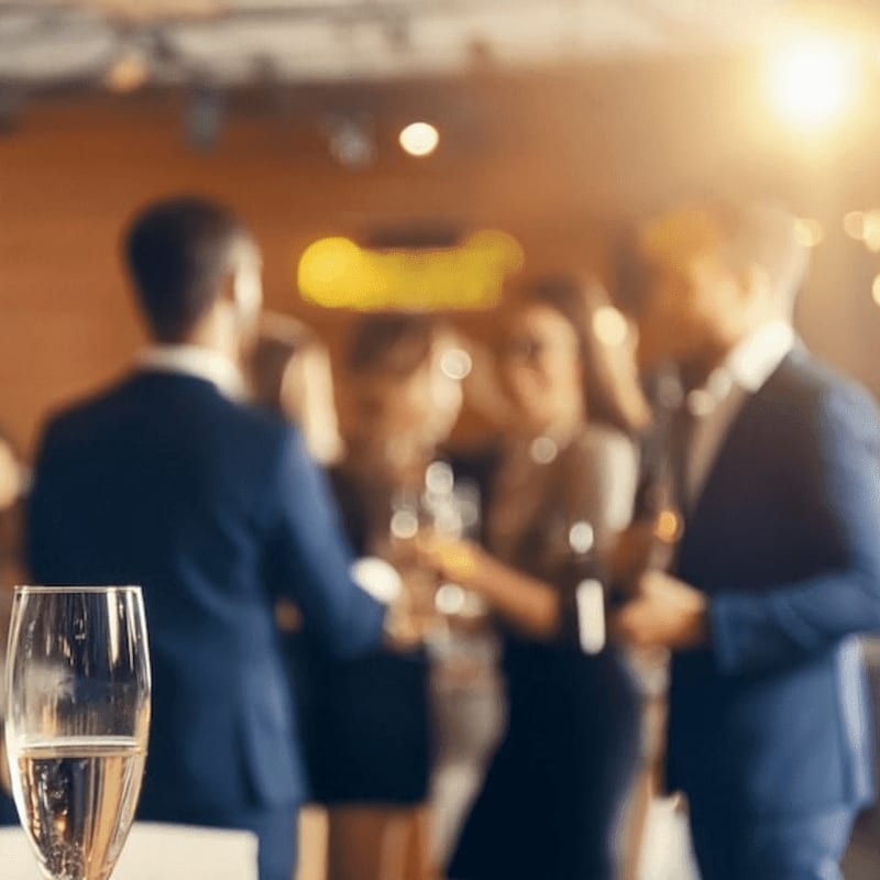 A champagne flute in the foreground is set against a blurred background of elegantly dressed people at a formal gathering.