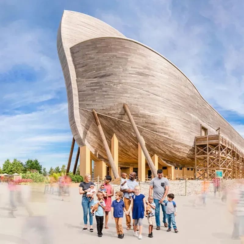 A group of families walks towards a massive wooden ark, with blurred figures and lush greenery in the background under a bright blue sky.