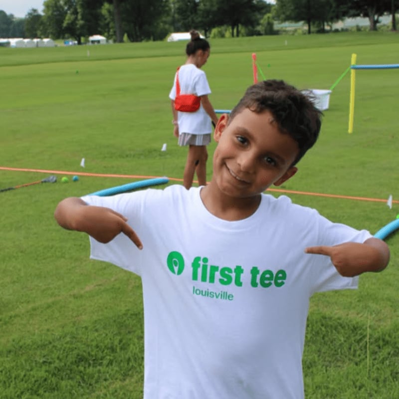 A young boy in a white t-shirt points to the "First Tee Louisville" logo while a girl stands in the background on a grassy golf course.